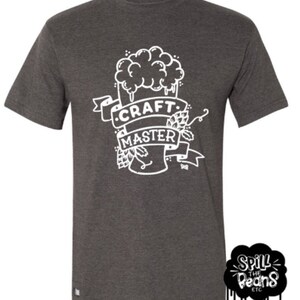 May include: A dark gray t-shirt with a white graphic of a beer glass with hops and the text "CRAFT MASTER". The graphic is in a vintage style. The t-shirt is from the brand "Spill the Beans ETC."
