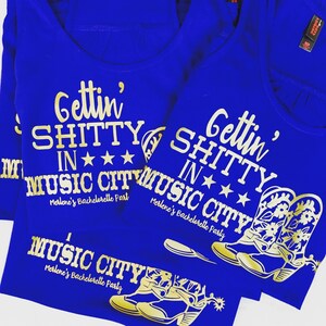 Op de afbeelding: Drie blauwe T-shirts met gouden glittertekst die "Gettin' Shitty in Music City" zegt en een gouden glitter-laarsontwerp. De tekst "Merlene's Bachelorette Party" is ook op de shirts opgenomen.