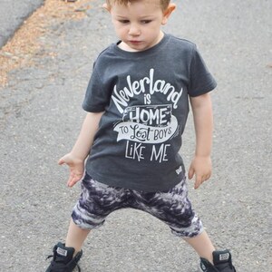 May include: A young boy wearing a dark grey t-shirt with the text "Neverland is home to lost boys like me" in white. He is wearing black and white patterned shorts and black high-top sneakers.