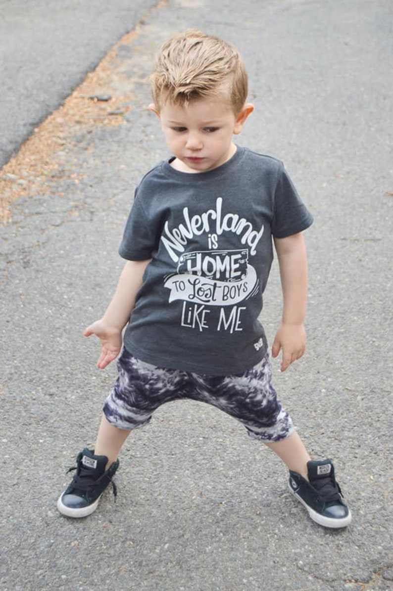 May include: A young boy wearing a dark grey t-shirt with the text "Neverland is home to lost boys like me" in white. He is wearing black and white patterned shorts and black high-top sneakers.