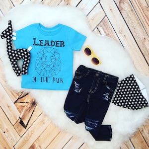 May include: A light blue t-shirt with a black geometric lion design and the text "Leader of the Pack". The shirt is paired with dark blue distressed jeans and a black and white patterned bandana.