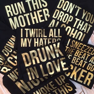 May include: A stack of black t-shirts with gold lettering. The shirts have various phrases printed on them, including "Run This Mother", "Don't You Drop That Alcohol", "I Twirl All My Haters", "Drunk In Love", "I Sneezed The Beat And The Beat Got Coker", and "Woke Up This Bitch Down".