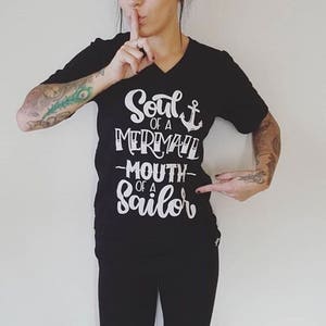 Puede incluir: Camiseta negra de cuello en V con texto blanco que dice "Soul of a Mermaid -Mouth of a Sailor" con un símbolo de ancla.