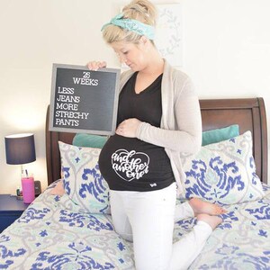 May include: A pregnant woman holding a letter board that reads "25 Weeks Less Jeans More Stretchy Pants". She is wearing a black shirt with the text "And Another One" on the front.