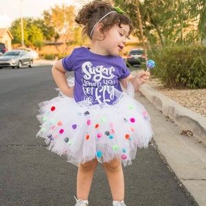 K&ouml;nnte beinhalten: Ein junges M&auml;dchen tr&auml;gt ein lila T-Shirt mit dem Text "Sugar, how'd you get so fly" und einen wei&szlig;en T&uuml;llrock mit bunten Bommeln. Sie h&auml;lt einen Lolli in ihrer rechten Hand.