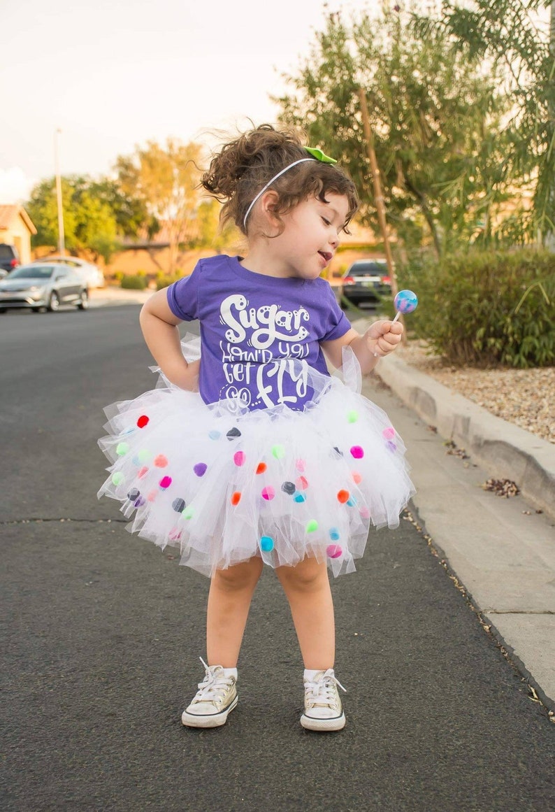 K&ouml;nnte beinhalten: Ein junges M&auml;dchen tr&auml;gt ein lila T-Shirt mit dem Text "Sugar, how'd you get so fly" und einen wei&szlig;en T&uuml;llrock mit bunten Bommeln. Sie h&auml;lt einen Lolli in ihrer rechten Hand.