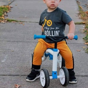 May include: A young child wearing a gray t-shirt with the text "They See Me Rollin'" and a yellow baseball cap rides a white and blue balance bike with black wheels.