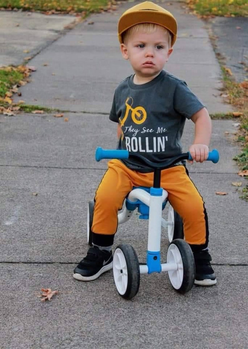 May include: A young child wearing a gray t-shirt with the text "They See Me Rollin'" and a yellow baseball cap rides a white and blue balance bike with black wheels.