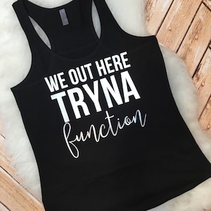 May include: Black racerback tank top with the text "WE OUT HERE TRYNA function" in white and silver lettering. The tank top is made of a soft material and is displayed on a white fluffy surface.