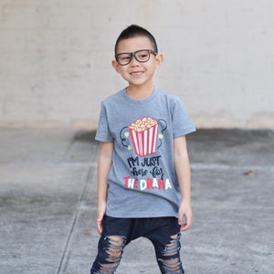 May include: A gray t-shirt with a red and white popcorn bucket graphic and the text "I'm just here for the drama".
