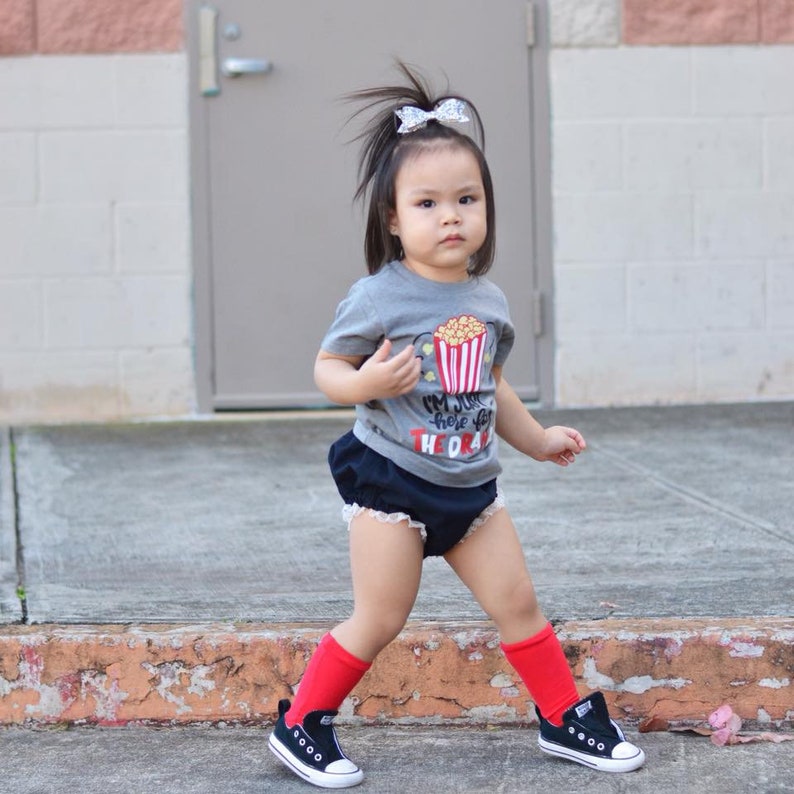 May include: A young girl wearing a grey t-shirt with a popcorn graphic and the text "I'm just here for the drama". She is also wearing black shorts, red socks, and black sneakers.