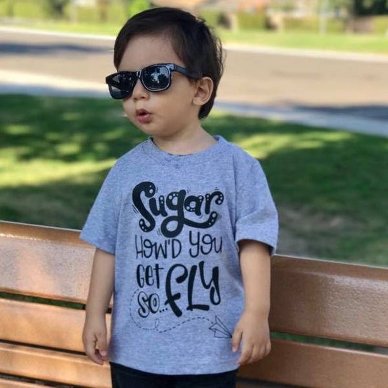 K&ouml;nnte beinhalten: Ein kleines Kind tr&auml;gt ein graues T-Shirt mit dem Text "Sugar How'd you get so... Fly" in schwarzen Buchstaben. Das Kind tr&auml;gt eine schwarze Sonnenbrille und sitzt auf einer Holzbank.