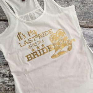 Puede incluir: Camiseta blanca de tirantes con texto en purpurina dorada que dice "It's My Last Ride Before I'm A Bride" con un par de botas vaqueras de purpurina dorada.