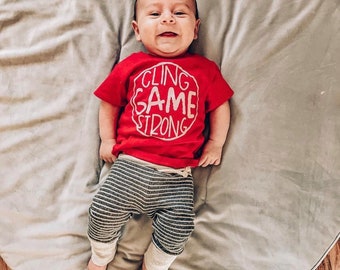 May include: A baby wearing a red shirt with the text "CLING GAME STRONG" in white. The baby is lying on a gray round mat.
