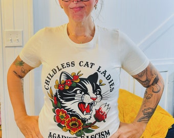 May include: A white t-shirt with a black and white cat illustration wearing a flower crown. The text "Childless Cat Ladies Against Fascism" is printed in a circle around the cat.