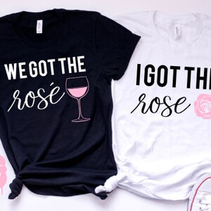 May include: Two black and white t-shirts with the text "We got the ros&eacute;" and a glass of ros&eacute; wine on one shirt and "I got the rose" with a pink rose on the other shirt.
