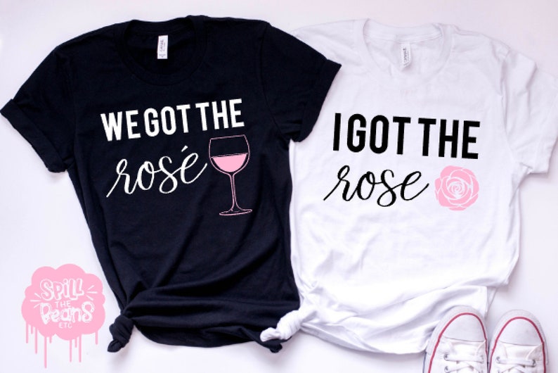 May include: Two black and white t-shirts with the text "We got the ros&eacute;" and a glass of ros&eacute; wine on one shirt and "I got the rose" with a pink rose on the other shirt.