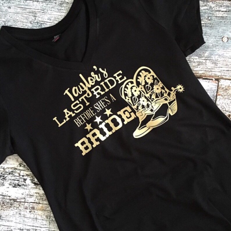 K&ouml;nnte beinhalten: Schwarzes T-Shirt mit goldener Aufschrift "Taylor's Last Ride Before She's A Bride" und einem Paar goldenen Cowboystiefeln.