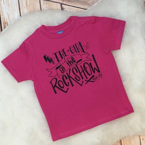 May include: A vibrant pink t-shirt featuring the black text "THE GIRL at the ROCKSHOW". The design includes lightning bolts and a banner. The t-shirt has short sleeves and a crew neck.