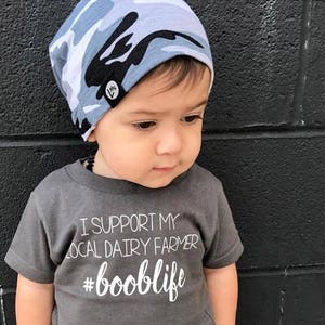 May include: A young child wearing a gray t-shirt with the text "I support my local dairy farmer #booblife" and a blue and white camouflage beanie.