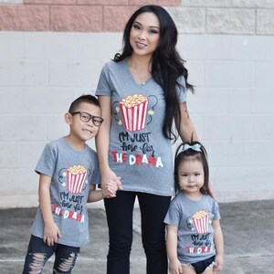 May include: A family of three wearing matching gray t-shirts with a popcorn bucket graphic and the text "I'm just here for the drama".