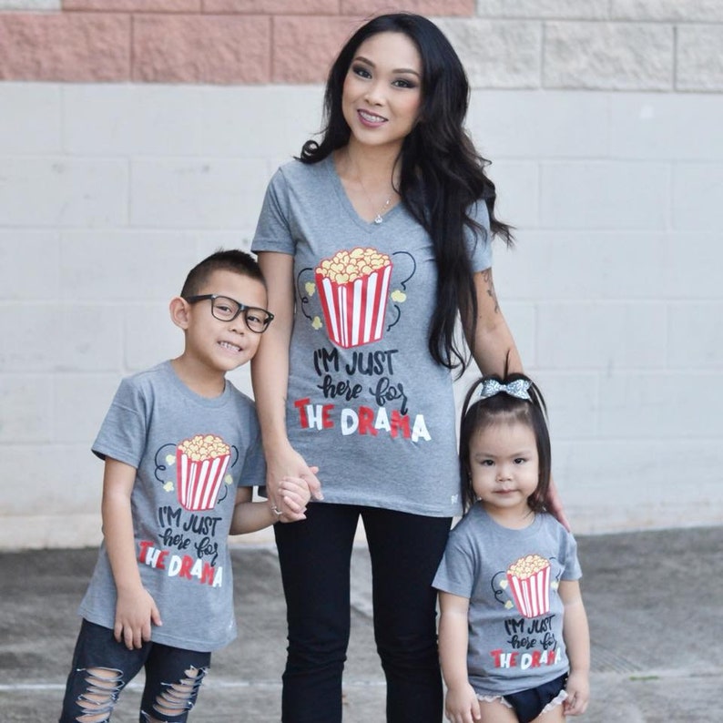 May include: A family of three wearing matching gray t-shirts with a popcorn bucket graphic and the text "I'm just here for the drama".