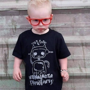 May include: A young child wearing a black t-shirt with a white robot graphic and the text "Intergalactic Planetary". The child is wearing red glasses and a silver bracelet.