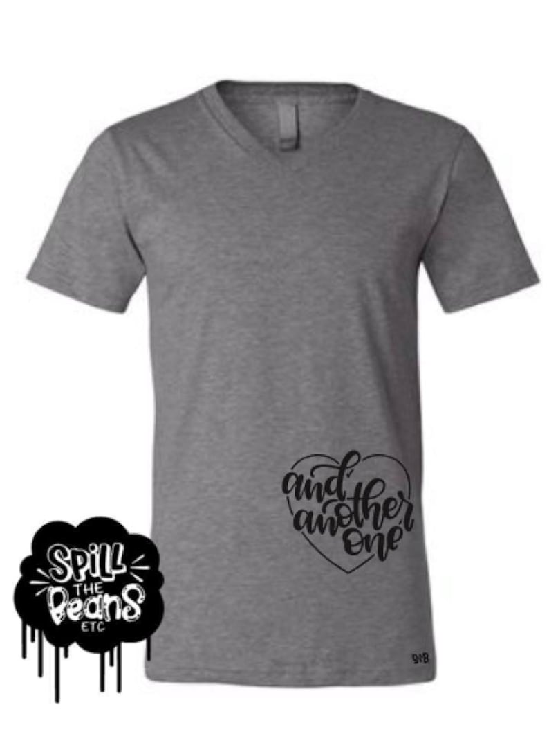 May include: A heather grey V-neck t-shirt with the text "and another one" in a black heart shape on the right side of the shirt.