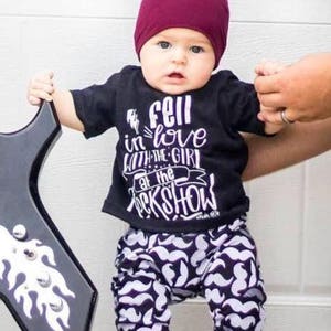 May include: A baby wearing a black t-shirt with the text "fell in love with the girl at the rock show" and black and white patterned trousers with a moustache design. The baby is wearing a burgundy beanie.