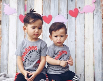 May include: Two young boys wearing grey t-shirts with a dinosaur graphic and the text "Love Bites". The boys are sitting on a red bench in front of a wooden fence decorated with paper hearts and dinosaurs.