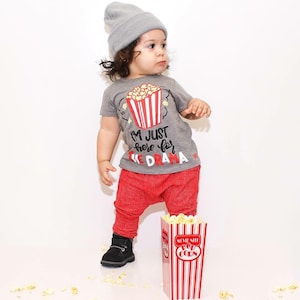 May include: A toddler wearing a gray t-shirt with the text "I'm just here for the drama" and a red popcorn bucket graphic. The toddler is also wearing red pants and a gray beanie. A red and white striped popcorn box is in the foreground.