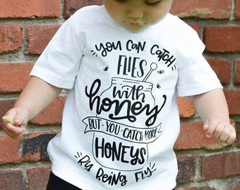 May include: A white t-shirt with black text that reads "You can catch flies with honey but you catch more honeys by being fly". The shirt is being worn by a child wearing black ripped jeans and a white baseball cap with a black star.