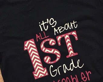 Może przedstawiać: Czarny T-shirt z białym tekstem, który brzmi "it's all about 1st grade mrs. Koehler". Cyfra "1" jest różowa i biała z wzorem w chevron.