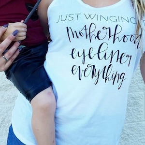 Pode incluir: Camisola branca de alças com o texto "Just Winging It Motherhood Eyeliner Everything" em escrita cursiva preta.