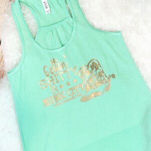 Op de afbeelding: Mintgroen tanktop met gouden glittertekst die "Gettin' Shitty in Music City" zegt met een paar gouden glittercowboylaarzen.
