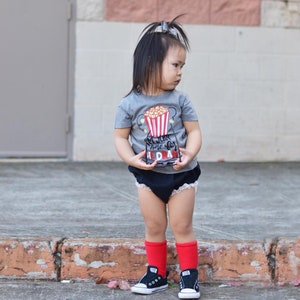 May include: A young child wearing a grey t-shirt with a popcorn bucket graphic and the text "I'm just here for the drama". The child is also wearing black bloomers, red socks, and black sneakers.