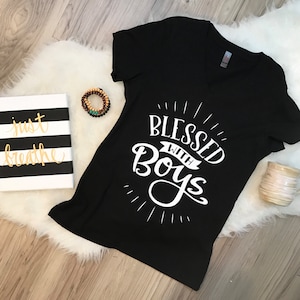 Puede incluir: Camiseta negra de cuello en V con el texto "Blessed with Boys" en letras blancas. El texto está en una fuente estilizada con una pancarta debajo de la palabra "Boys".
