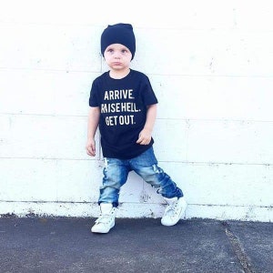 May include: A young child wearing a black t-shirt with the text "ARRIVE. RAISE HELL. GET OUT." printed in white, blue jeans, white sneakers, and a black beanie.