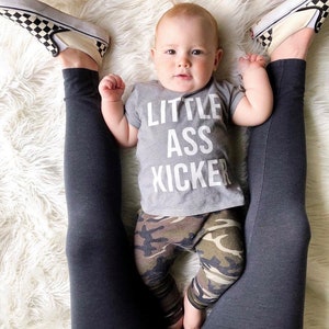 Puede incluir: Un bebé con una camiseta gris con el texto "Little Ass Kicker" y pantalones de camuflaje. El bebé está acostado en una alfombra blanca y esponjosa con un par de leggings negros a cada lado del bebé.