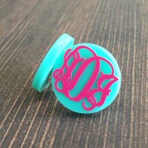 May include: A turquoise blue round disc with a pink monogram in the center. The monogram is a stylized letter "O" with a smaller "O" inside.