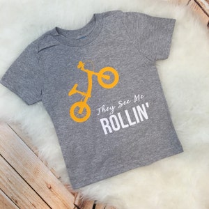 May include: A gray t-shirt with a yellow silhouette of a child's bike and the text "They See Me Rollin'" in white.
