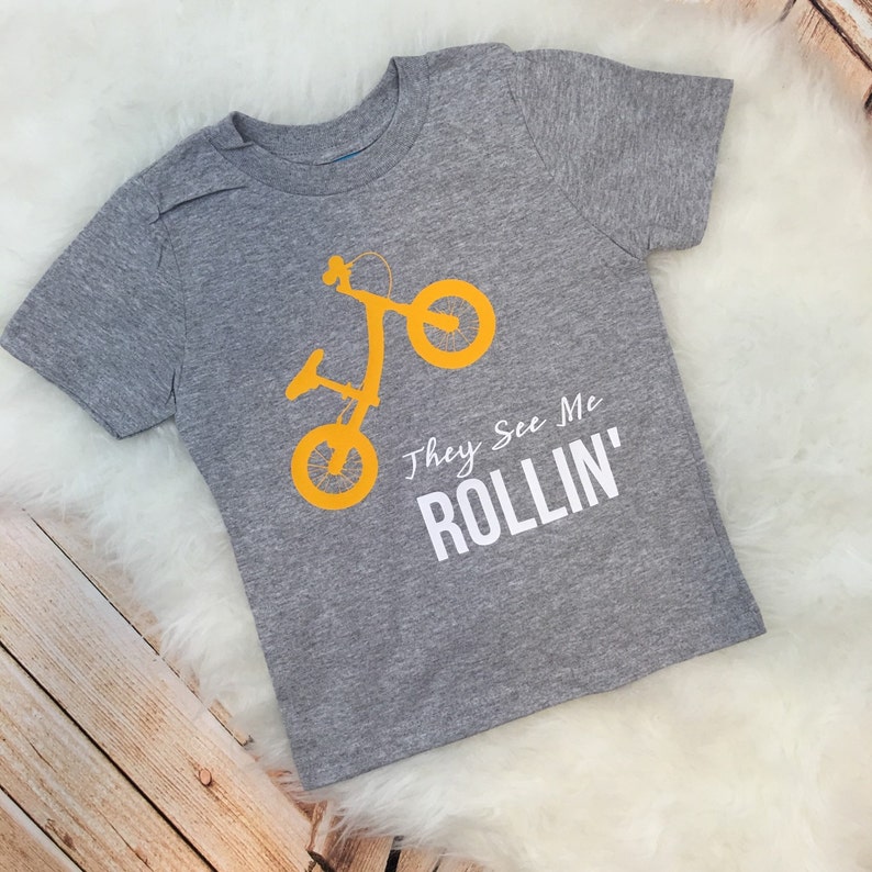 May include: A gray t-shirt with a yellow silhouette of a child's bike and the text "They See Me Rollin'" in white.