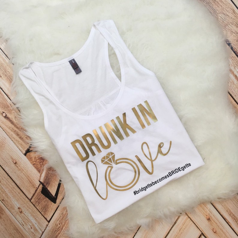 May include: White tank top with gold text that reads "Drunk in Love" with a gold ring and diamond graphic. The hashtag #bridgettebecomesBRIDEgette is printed below the text.