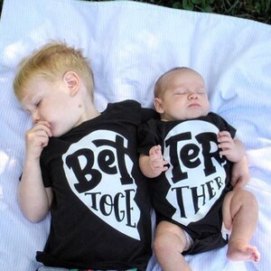 May include: Two babies wearing black t-shirts with a white heart design. The shirts say "Better Together" with each shirt having half of the heart and text. The babies are lying on a white blanket.