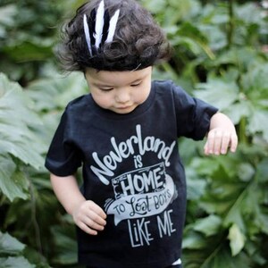 May include: A black t-shirt with a white graphic that says "Neverland is Home to Lost Boys Like Me". A young child is wearing the shirt and has a black and white feather headband in their hair.