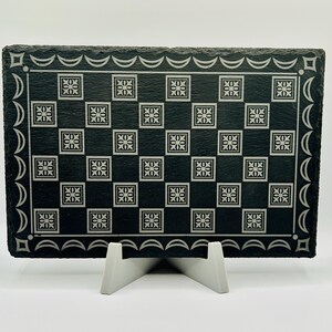 May include: A rectangular black slate serving board with a white floral checkerboard pattern. The board has a decorative white border and is supported by a white stand. The board is approximately 30 cm long and 20 cm wide.