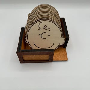 May include: A set of wooden coasters with a cartoon character's face, each with a simple line drawing. The coasters are stored in a wooden box with a dark brown frame and a light brown interior.