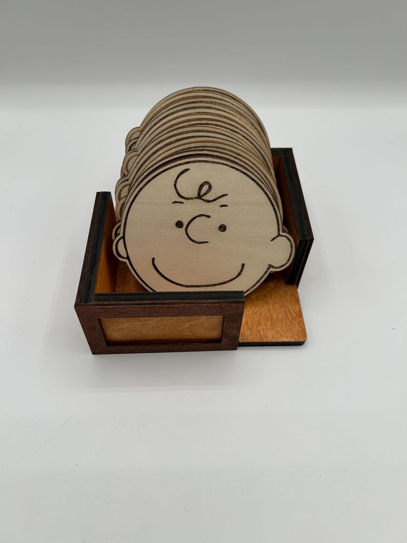 May include: A set of wooden coasters with a cartoon character's face, each with a simple line drawing. The coasters are stored in a wooden box with a dark brown frame and a light brown interior.