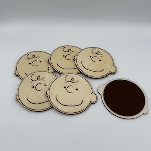 May include: A collection of six wooden coasters, each depicting a cartoon character's face. The coasters are light brown with dark brown outlines and details. One coaster has a dark brown felt base.