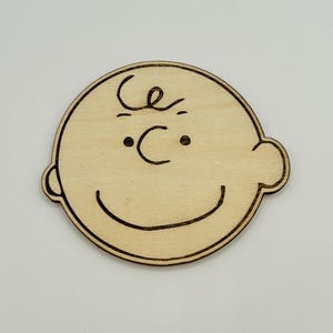 May include: A wooden cutout of a cartoon character's face. The design features a round head, small eyes, a curved smile, and a single curl of hair. The character's features are outlined in dark brown against the light wood.
